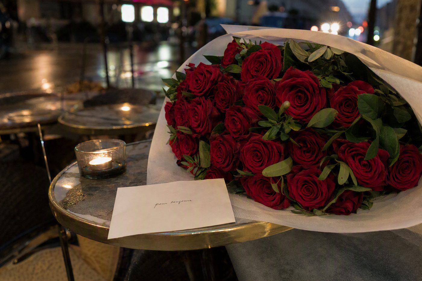 A bouquet of red roses beside a handwritten note on a Paris café table at night